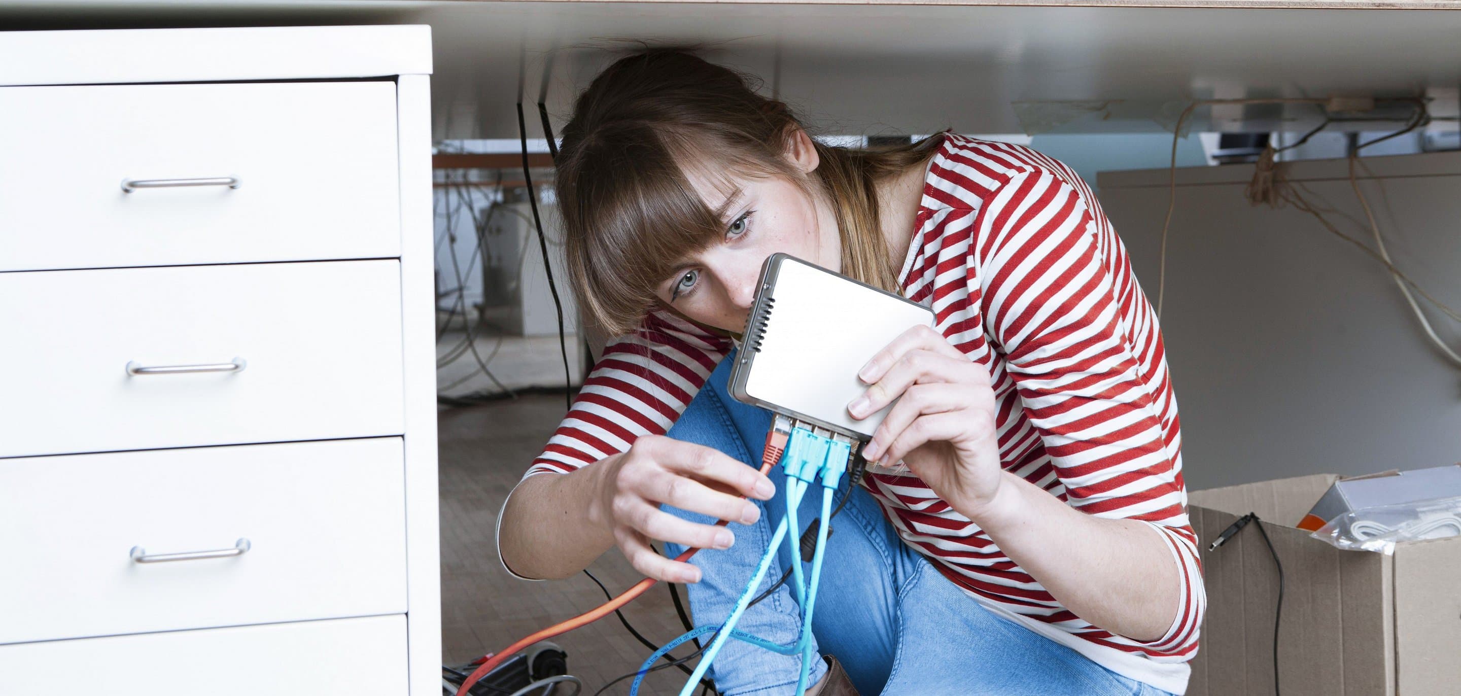 Junge Frau, die ein rot-weiß gestreiftes Oberteil trägt, sitzt unter ihrem Schreibtisch, hält einen Router in der Hand und zieht an einem Kabel.