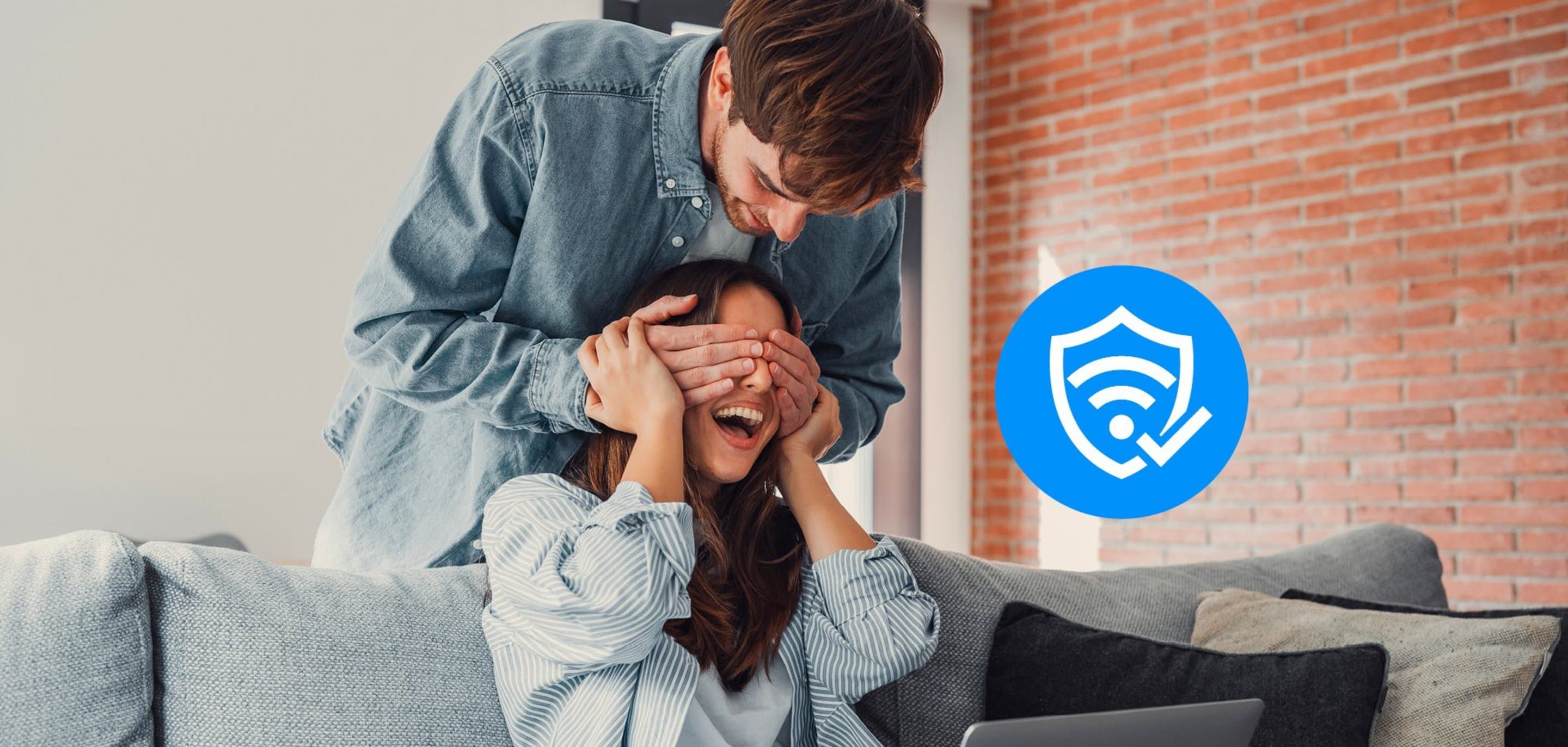 Junge Frau sitzt auf Couch und bekommt Augen von einem jungen Mann zugehalten. Neben dem Paar ist ein Sicherheits-Icon zu sehen.