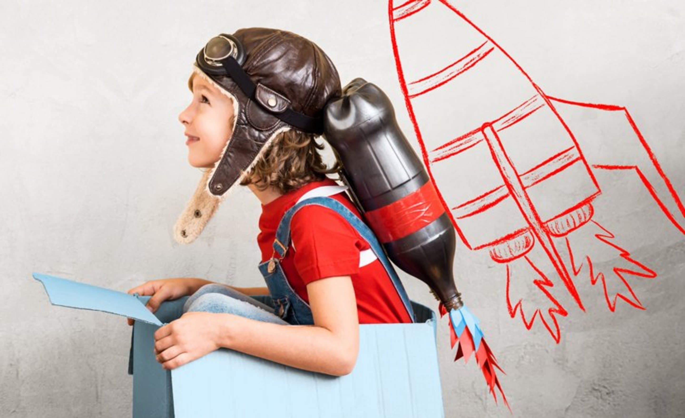 Kind sitzt vor einem grauen Hintergrund mit selbstgebasteltem Jetpack einem Karton; Szene symbolisiert Geschwindigkeit des Internets und erfolgreiches Durchstarten.