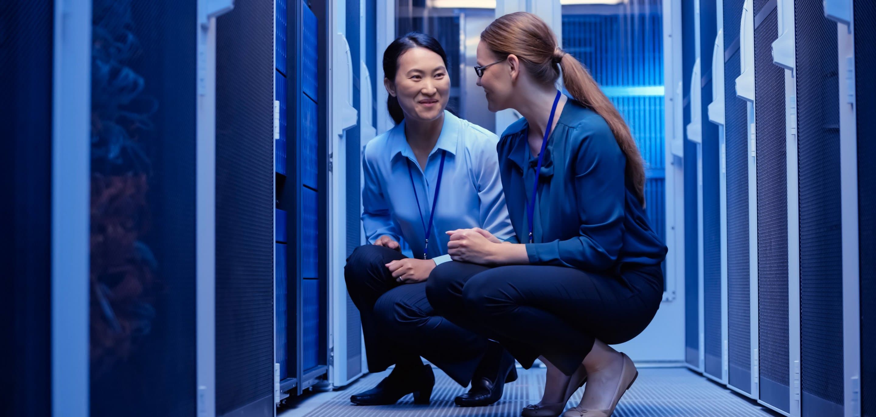Rechenzentrum Zwei Personen in Businesskleidung knien in einem blau beleuchteten Serverraum und unterhalten sich zwischen Serverracks.