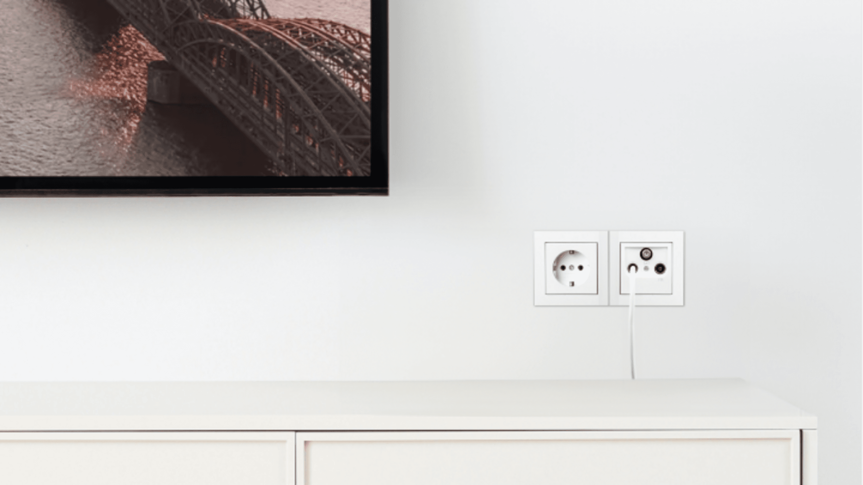 Kabel TV Fernseher zeigt eine Brücke über Wasser, darunter ein weißes Möbelstück, rechts eine Multimedia-Steckdose mit angeschlossenem Kabel.