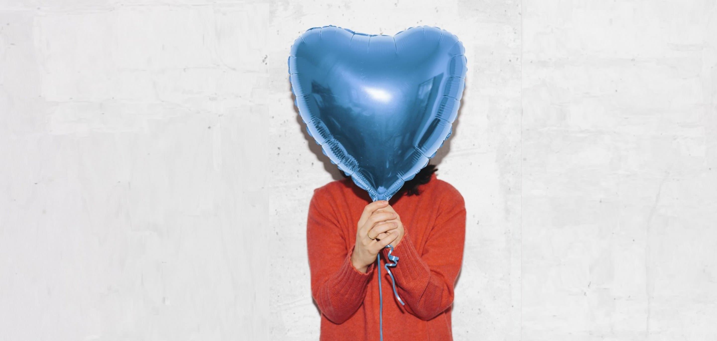 NetCologne Engagement Person in rotem Pullover hält einen großen blauen Herzballon vor einer hellen Betonwand.