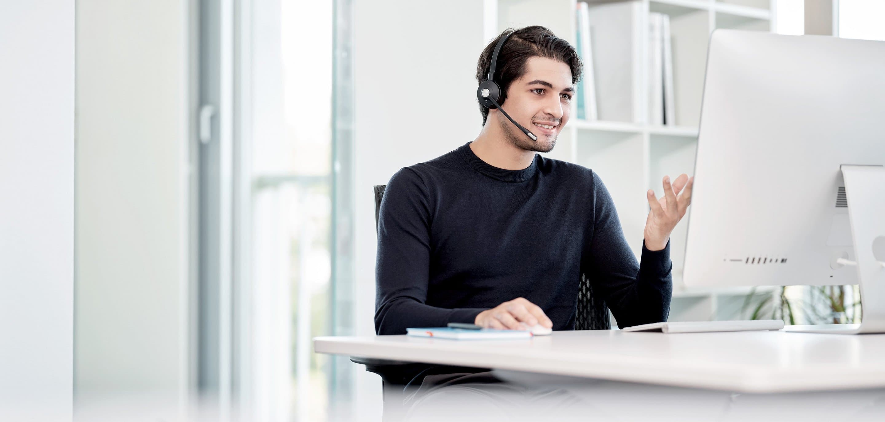 Digitaler Kundenkontakt Person mit Headset sitzt am Schreibtisch vor einem Monitor, offenbar in einem Video-Call, mit Büchern und Regalen im Hintergrund.