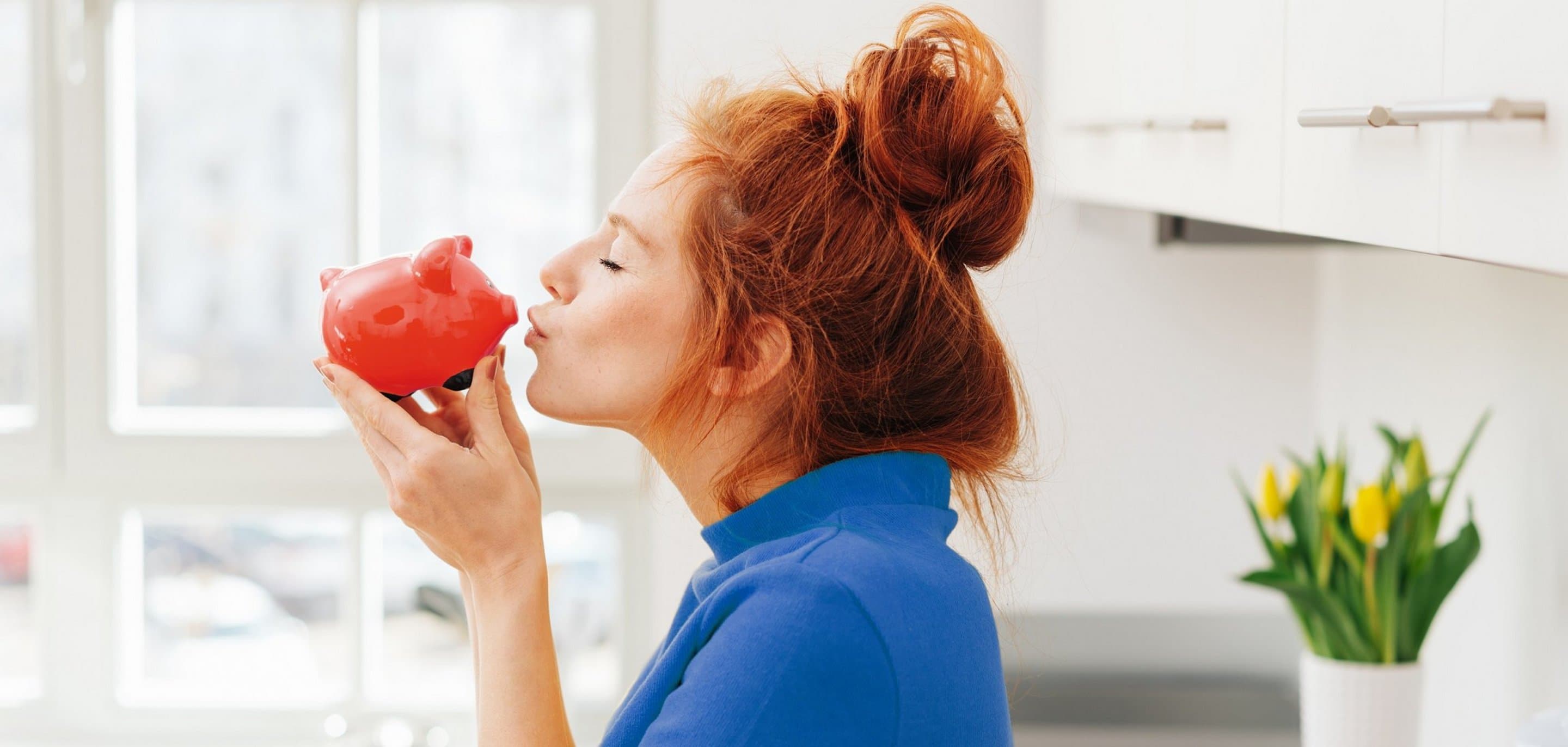 Rothaarige Frau mit Dutt küsst rotes Sparschwein.