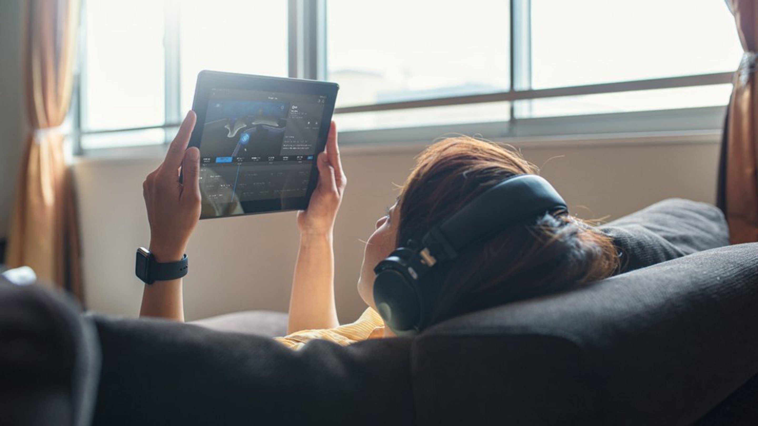 Couch von hinten auf der eine Frau liegt, die Kopfhörer trägt und Tablet in der Hand hält.